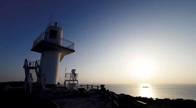断崖の上に立つ「大バエ灯台」が、水平線に沈み行く夕陽に照らし出される。