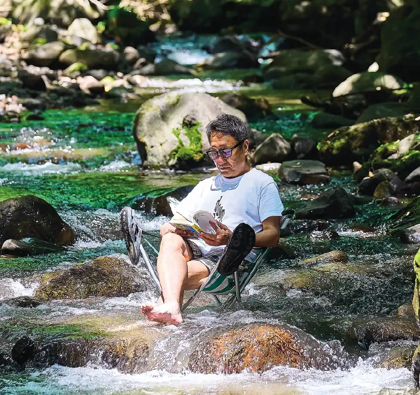 カートピア アウトドアチェアを川の中に設置し、足を水面に浸して読書をする酒井直人 | SUBARU