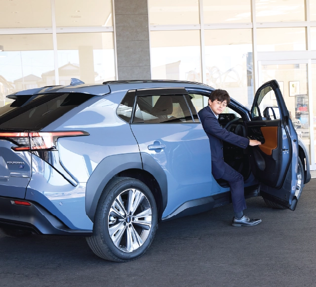 カートピア ソルテラ(ボディーカラーはハーバーミストグレーパール)の運転席ドアから片足を地面に下ろして半身を乗り出す、栃木スバルのセールス・清水さん。 | SUBARU