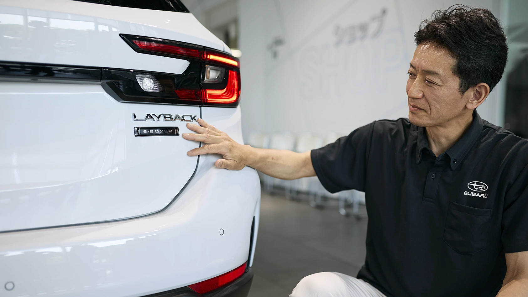 カートピア ストロングハイブリッド搭載のレイバックのリヤにあるエンプレムに手を添える、SUBARUデザイン部の源田哲朗。 | SUBARU