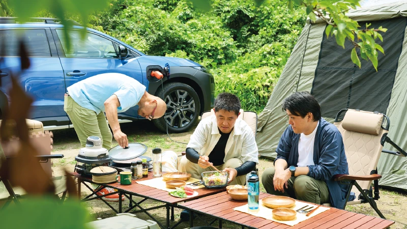 カートピア キャンプサイトのテーブルを囲み、電気調理家電を使って食事を愉しむ3人。 | SUBARU
