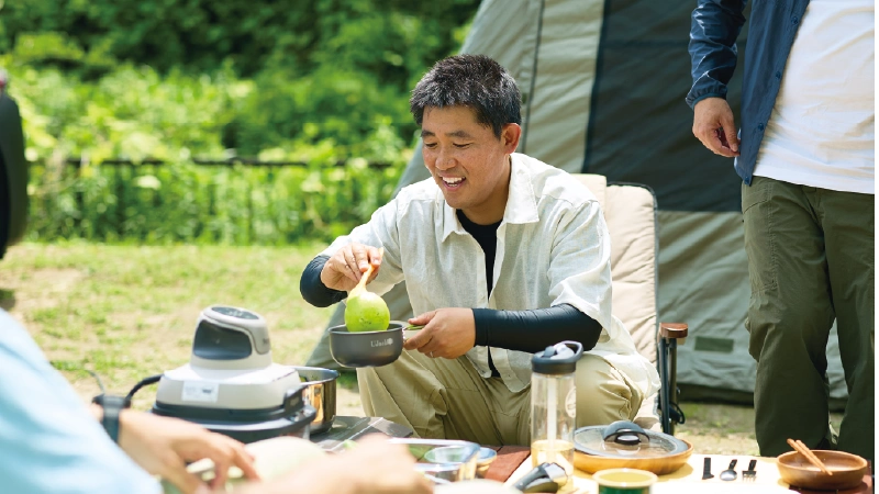 カートピア キャンプサイトのチェアに座り、食事を囲んでくつろぐ男性。 | SUBARU